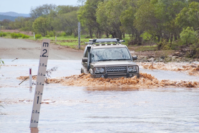 4x4 is slowly crossing flooded road next to a mark indecating waterlevel. Captured during rain season in Western Australia
